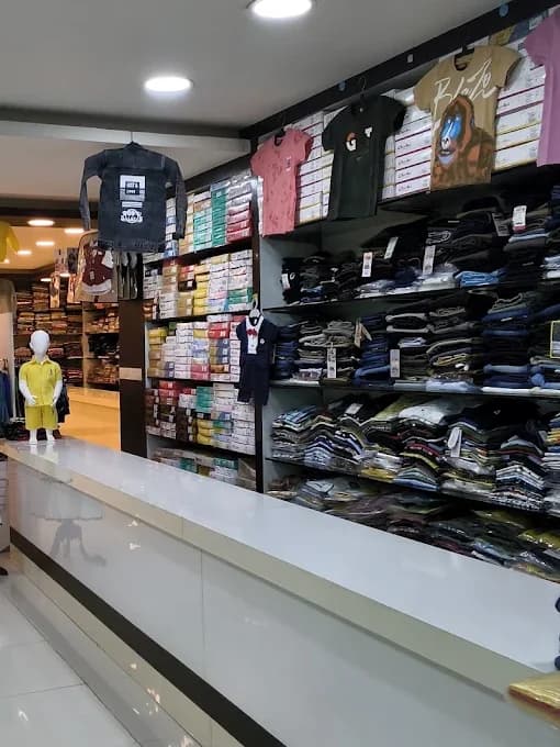 Kidswear at Laksha Fashion, Hosur: graphic T-shirts and a denim jacket on display above shelves of folded kids' clothing, with a child mannequin in yellow